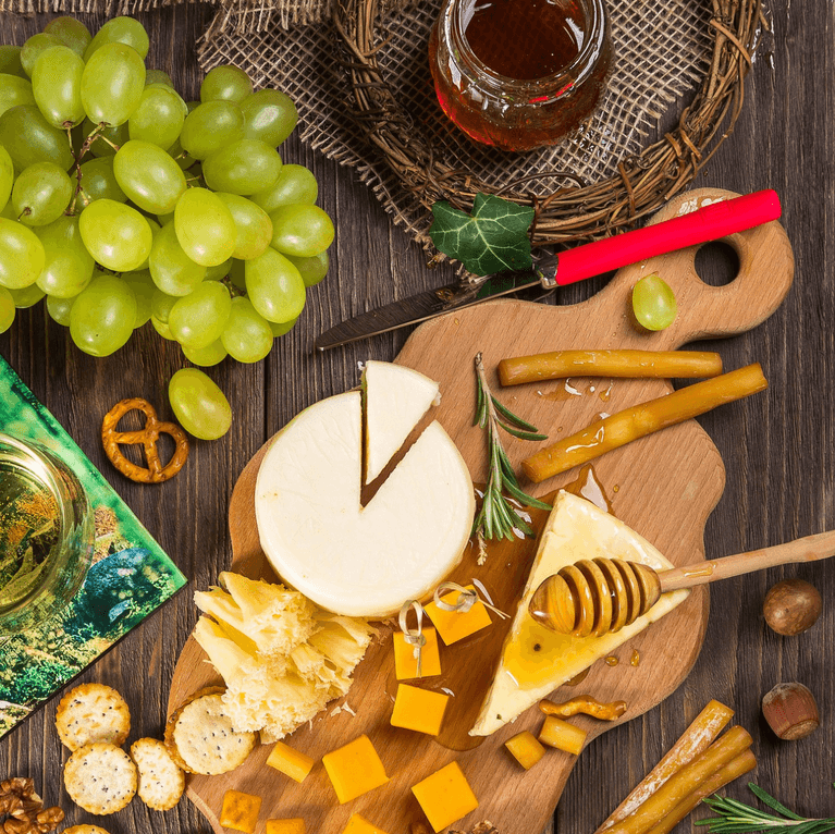 Overhead view of a cheese board with cheese, grapes, crackers, honey, and breadsticks on a wooden table.