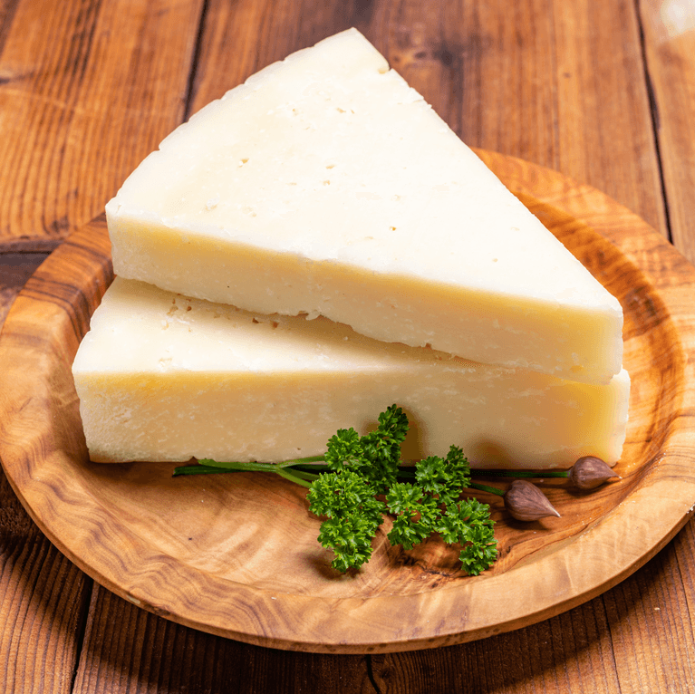 Two wedges of light-colored cheese, possibly Parmesan or Pecorino Romano, are stacked on a rustic wooden plate with sprigs of parsley and two garlic cloves. The image is on a wooden table against a blurred background.