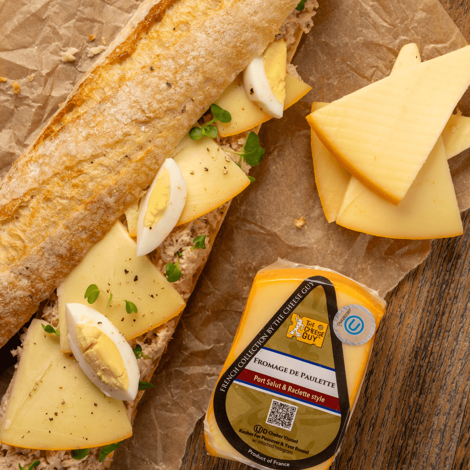 The Cheese Guy Fromage De Paulette cheese, wedge-shaped block, alongside a baguette sandwich with sliced Fromage De Paulette, hard-boiled eggs, and greens on parchment paper.