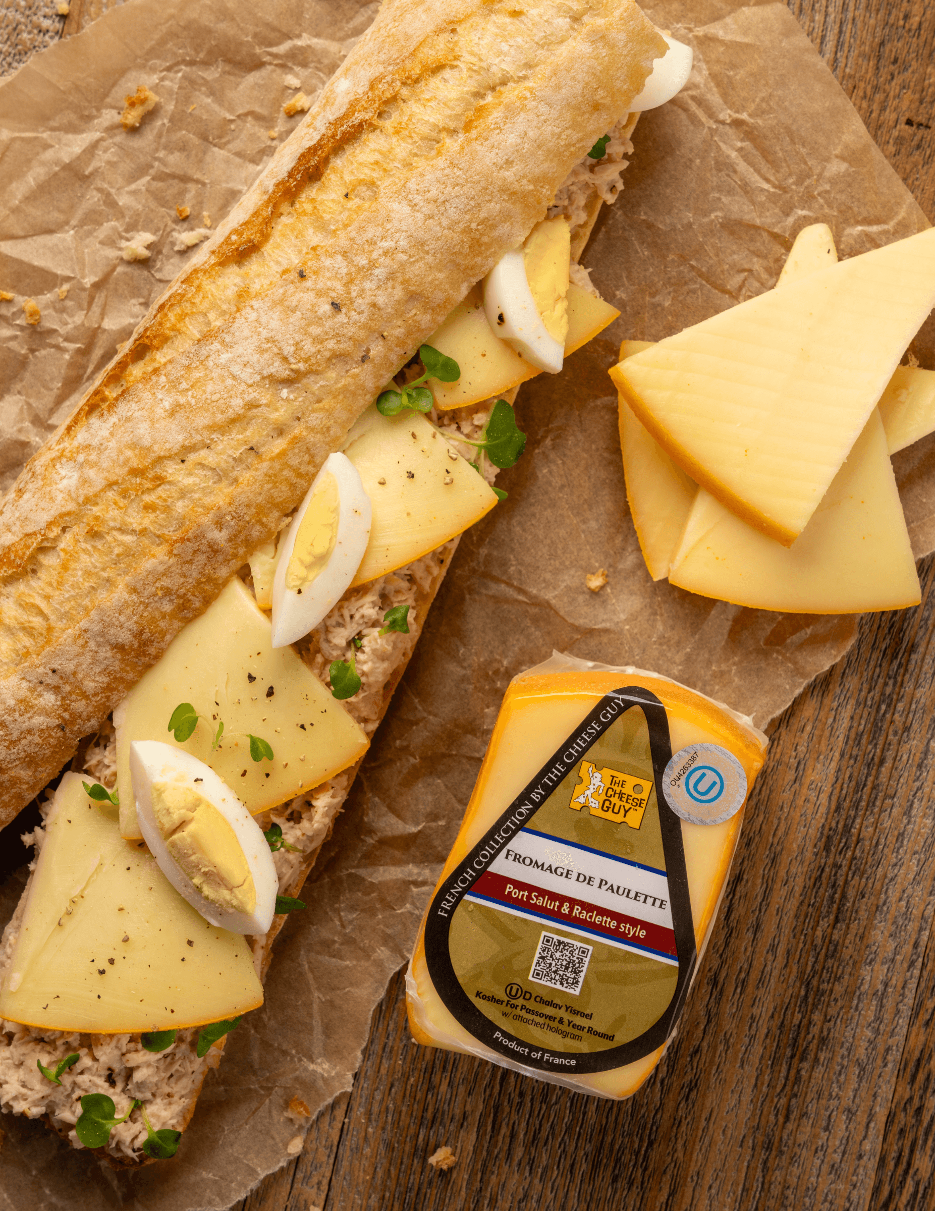 The Cheese Guy Fromage De Paulette cheese, wedge-shaped block, alongside a baguette sandwich with sliced Fromage De Paulette, hard-boiled eggs, and greens on parchment paper.