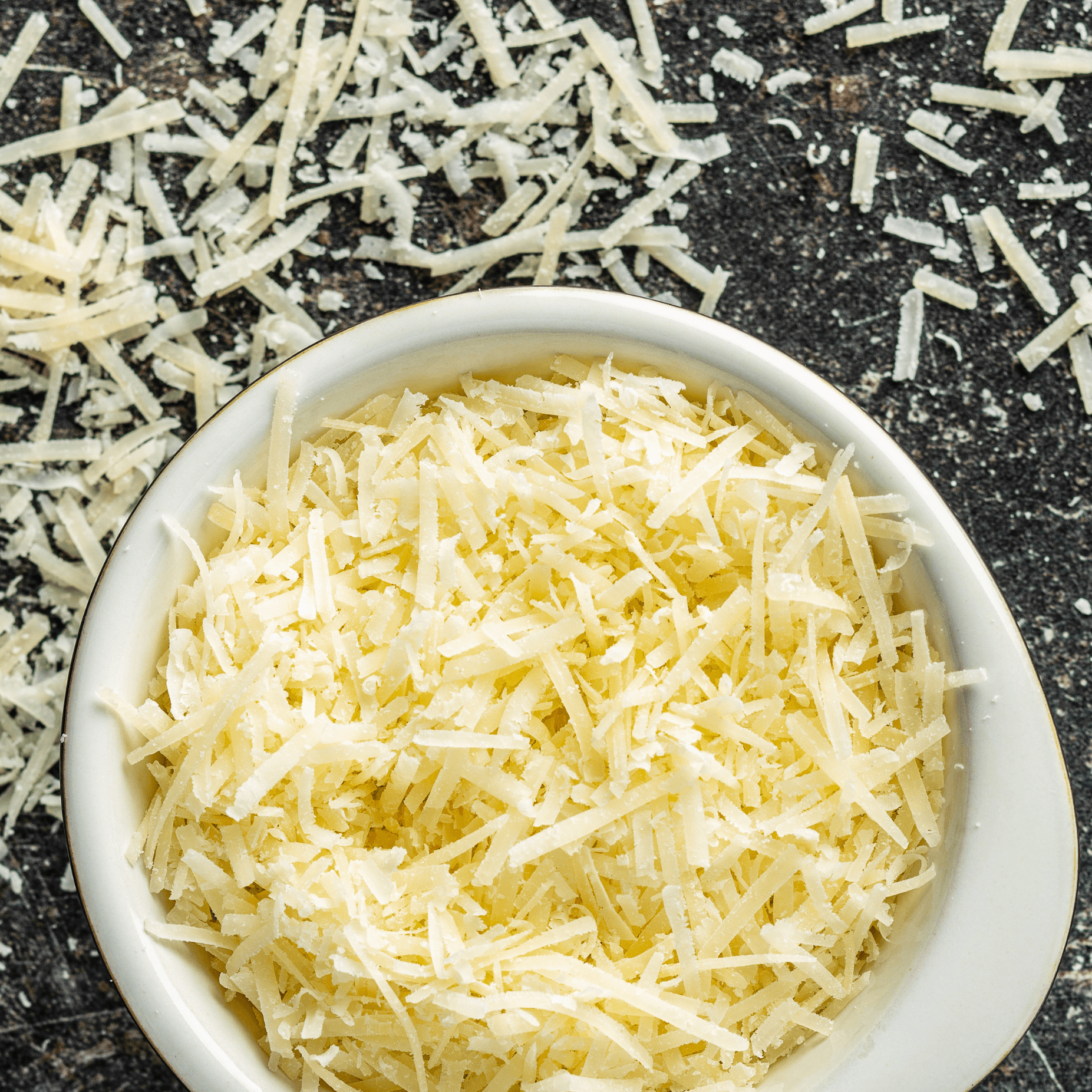 The top-down view is of a white bowl filled with grated Parmesan cheese, showing its light color and texture, with additional cheese shreds scattered on a dark, textured background.
