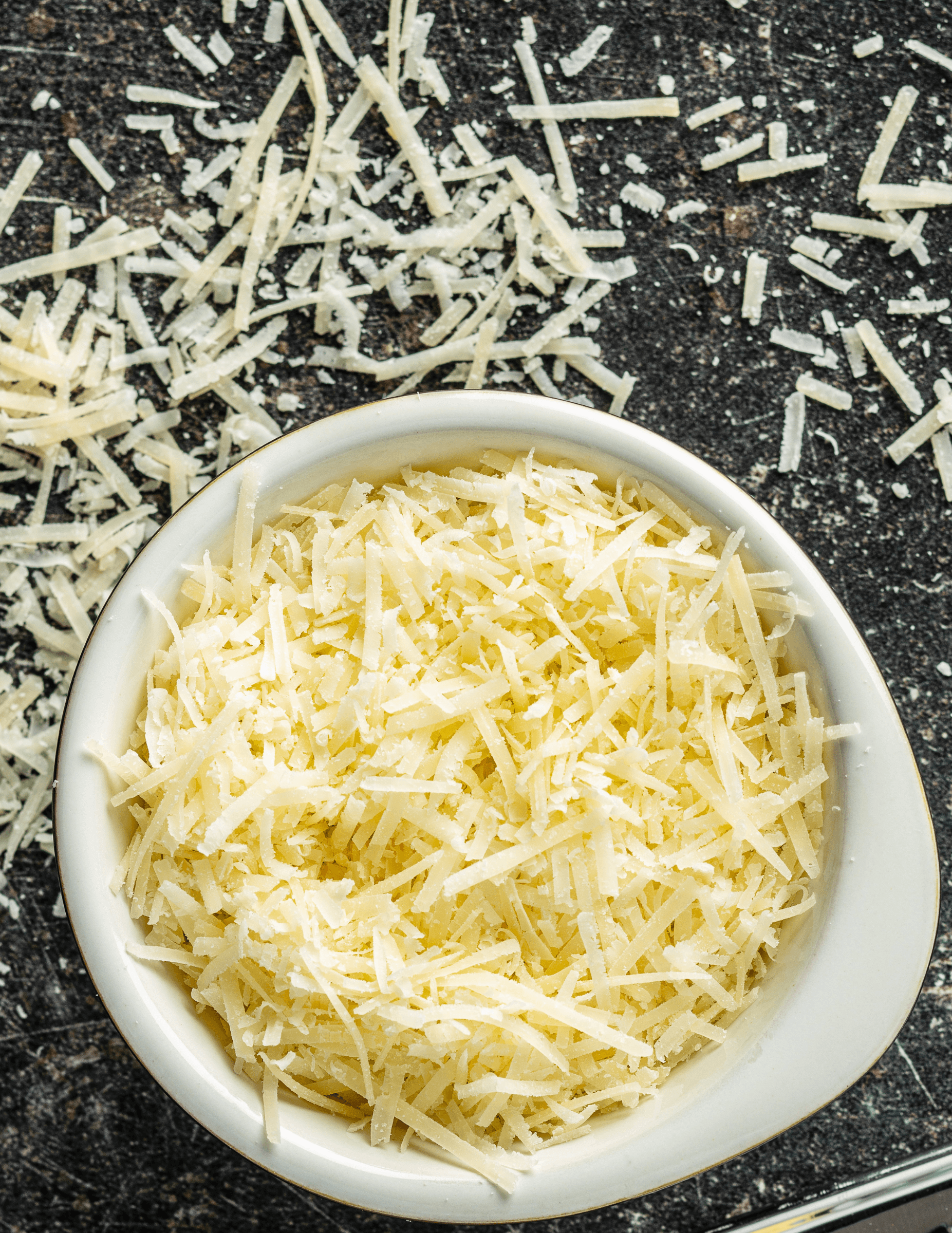 The top-down view is of a white bowl filled with grated Parmesan cheese, showing its light color and texture, with additional cheese shreds scattered on a dark, textured background.