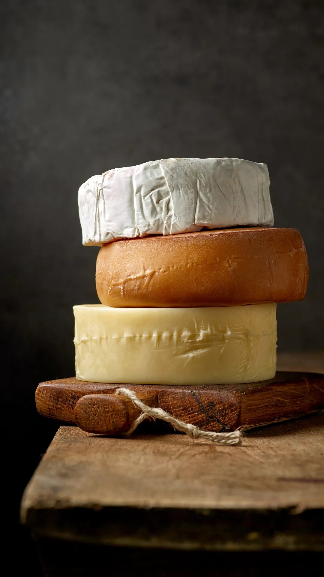 Three cheeses stacked on a wooden board against a dark background. Includes brie, gouda, and cheddar.