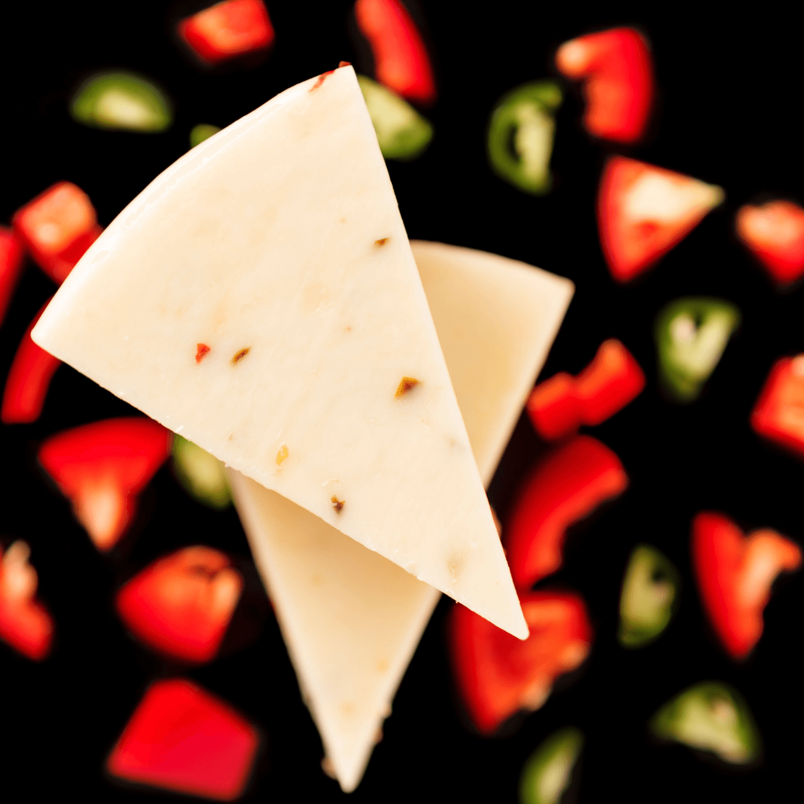 Two triangular slices of creamy white pepper jack cheese with visible flecks of red and green peppers arranged on a black surface scattered with diced red and green bell peppers.