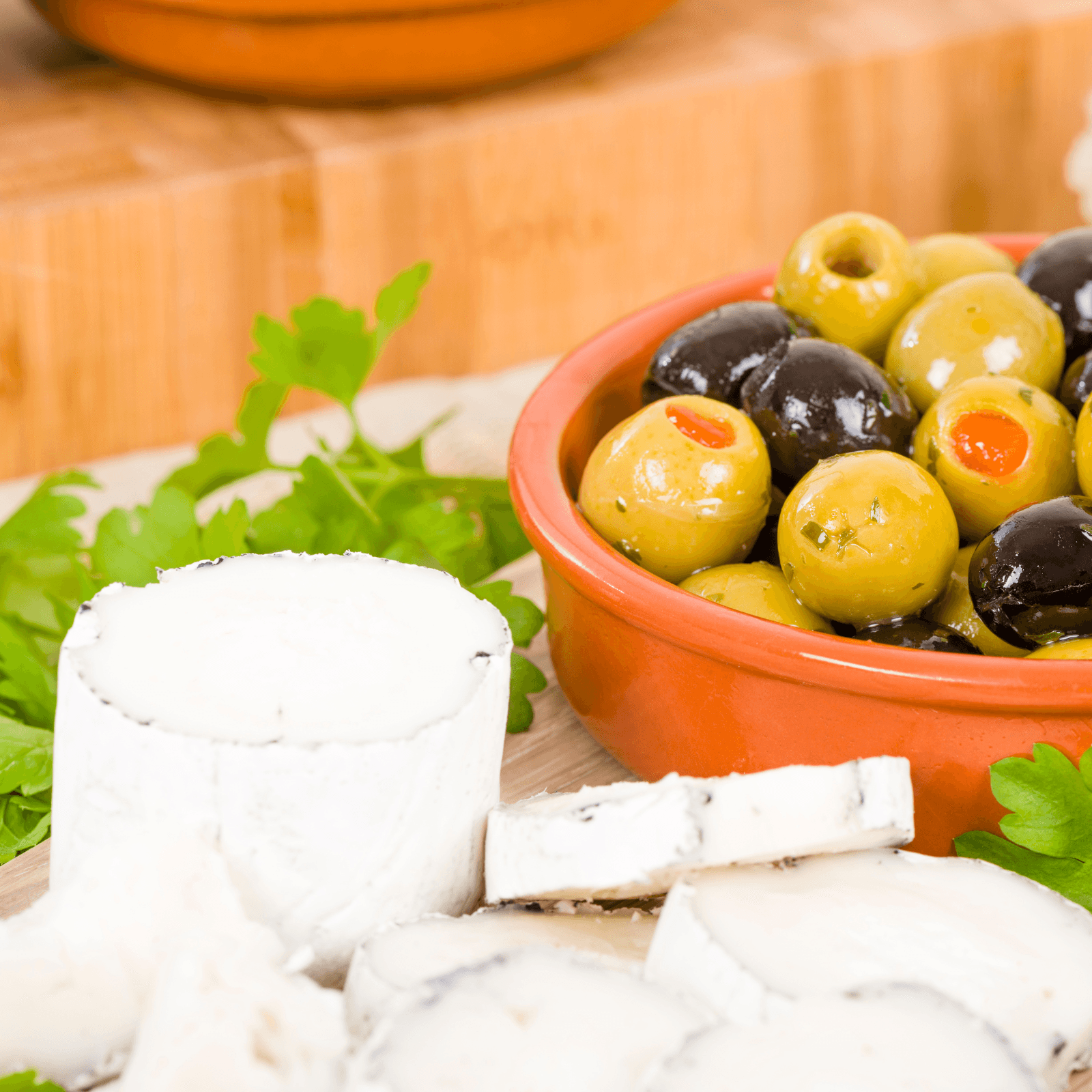 A close-up of a small orange bowl filled with mixed green and black olives, some stuffed with pimentos, next to a log of white goat cheese and several slices on a light-colored wooden board, garnished with fresh parsley leaves.