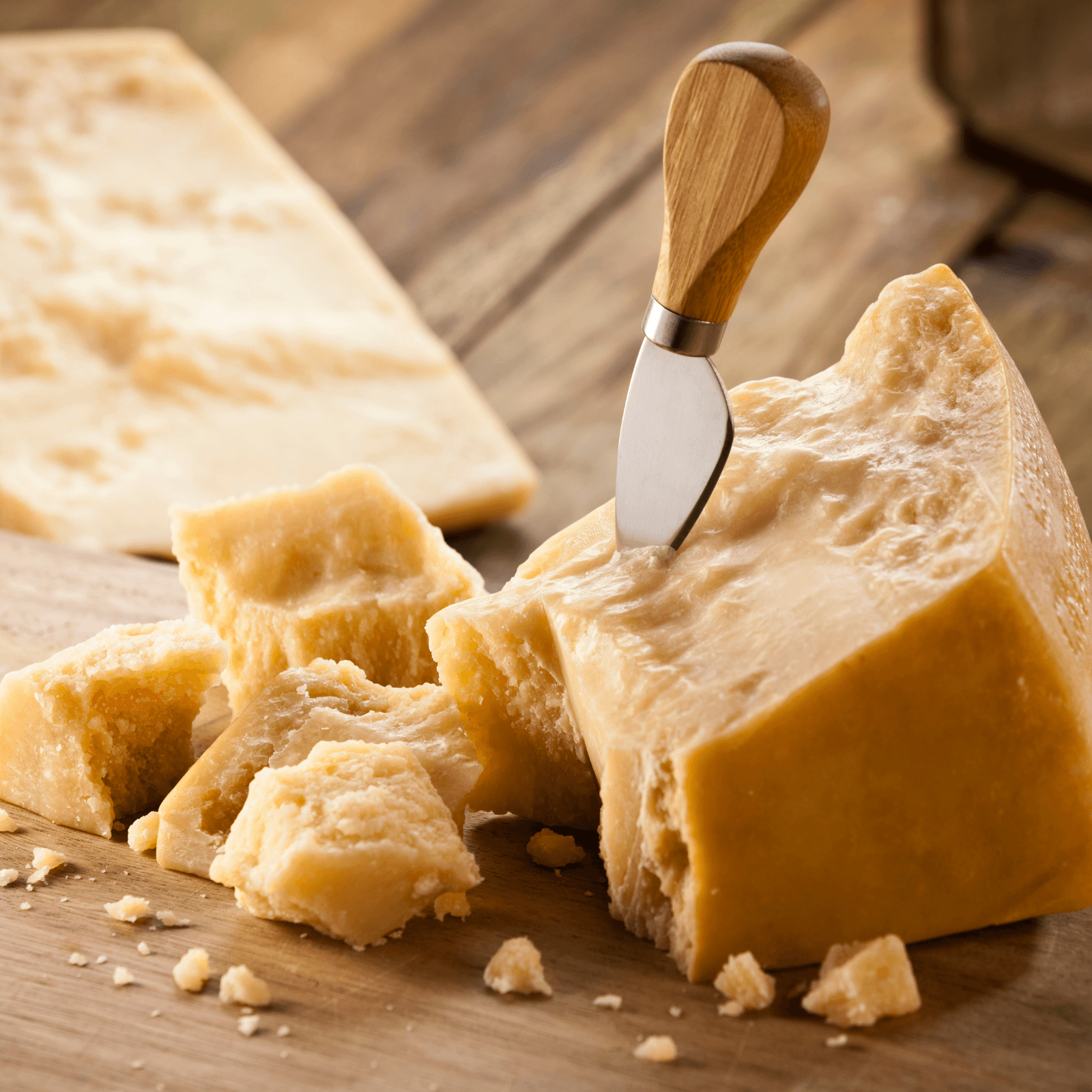 Close-up of aged Parmesan or Parmigiano Reggiano cheese chunks on a wooden surface, with a cheese knife inserted into a larger piece, showcasing the crumbly texture.