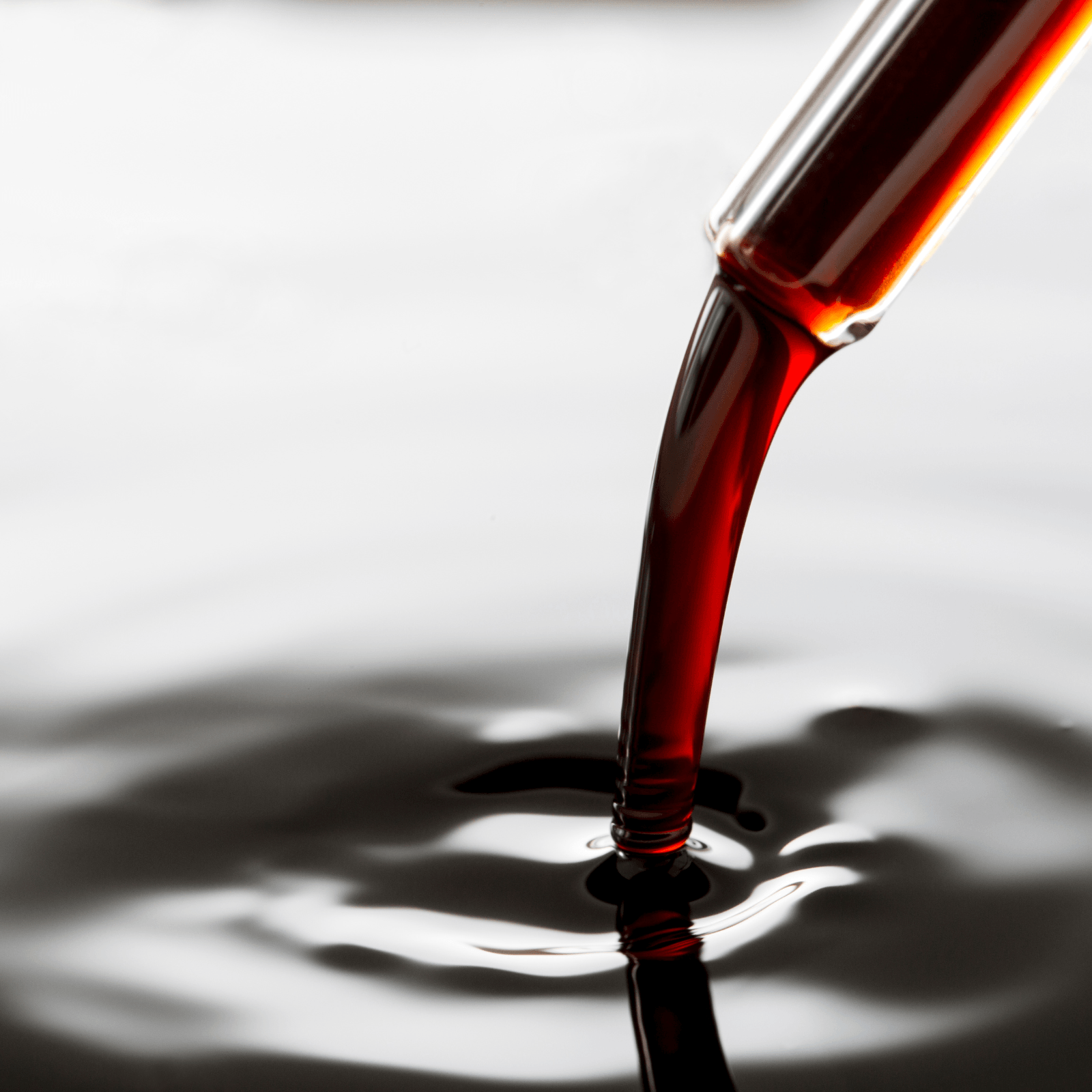 Close-up of a dark red liquid being dispensed from a clear glass pipette into a dark, rippled liquid, creating a small splash.