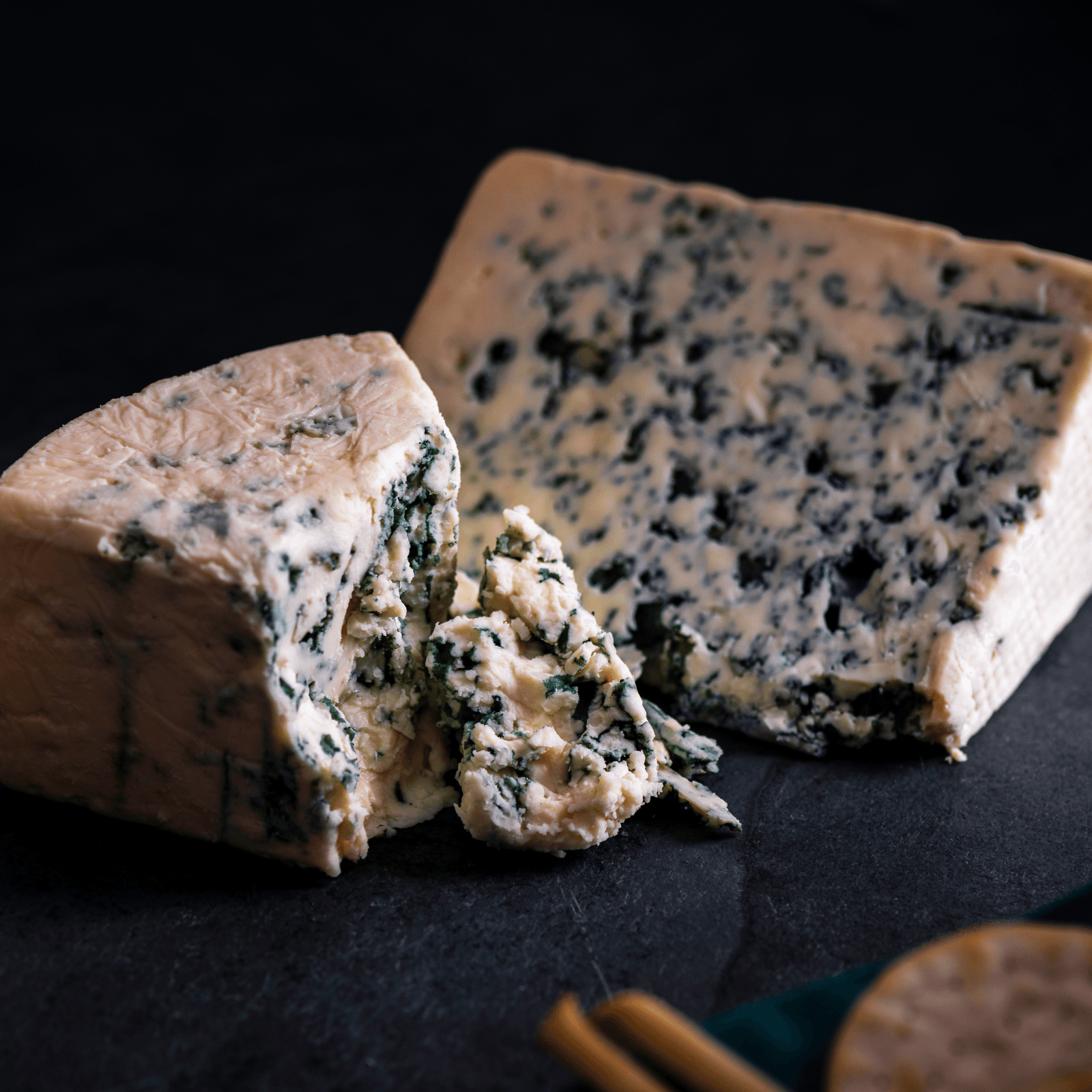 Image of two wedges of blue cheese with visible mold marbling, on a dark textured surface, with crackers in the corner.