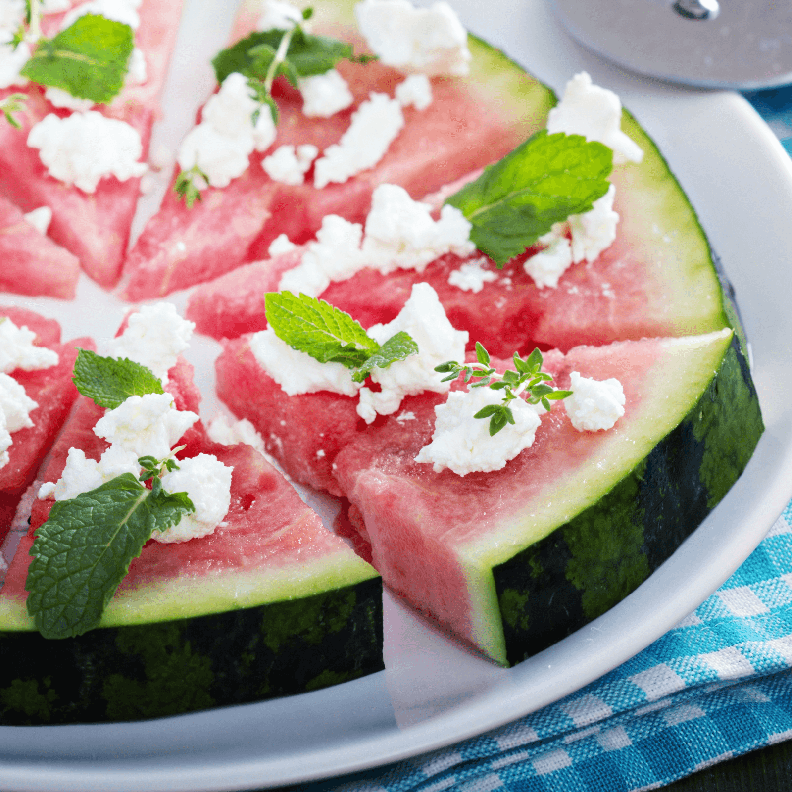 Image of watermelon pizza slices, arranged on a white plate, topped with crumbled feta cheese and fresh mint leaves, with a blue checkered napkin.