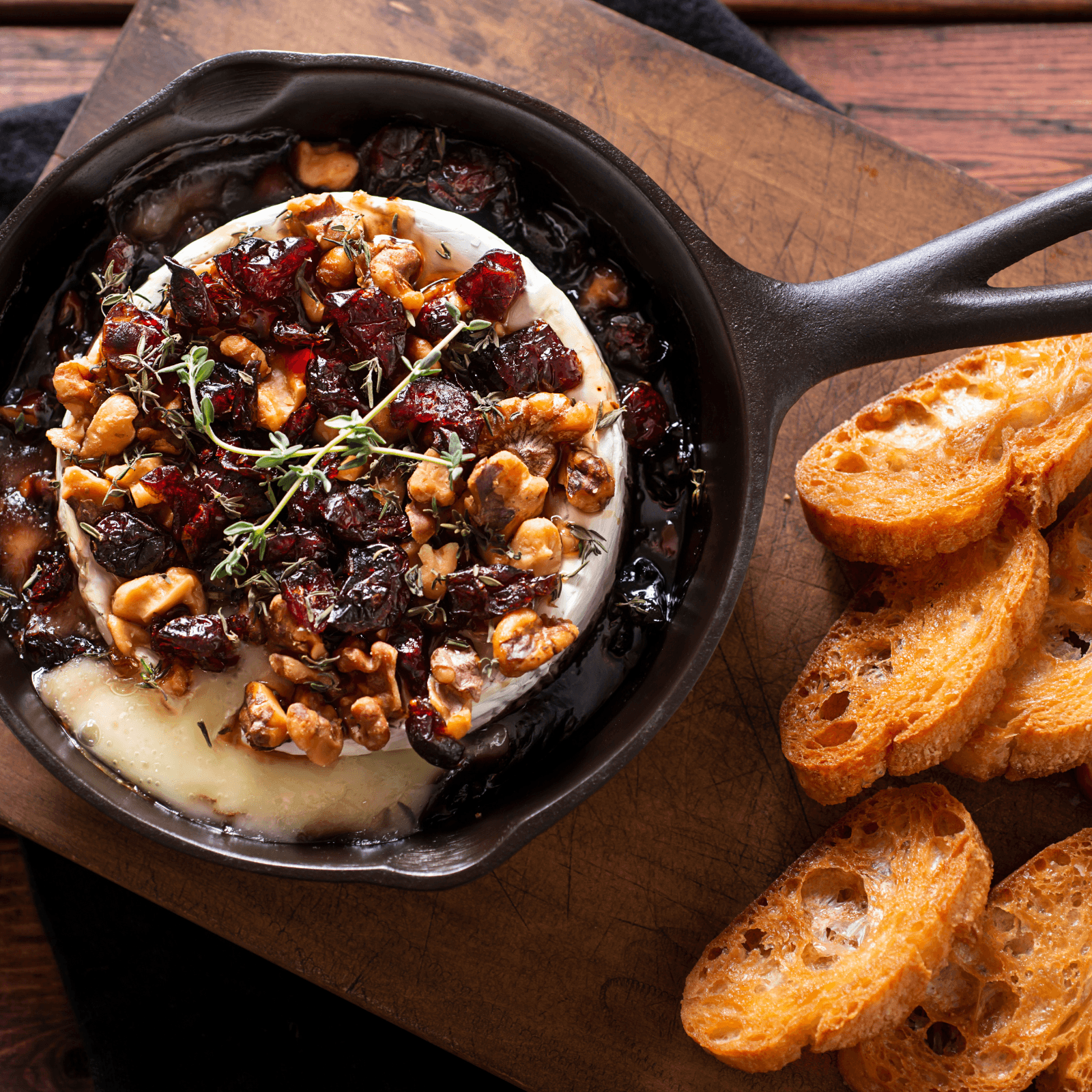 Close-up photo of baked brie in a small cast iron skillet. The brie is topped with cooked cranberries and walnuts, garnished with a sprig of thyme. A wooden board with slices of toasted bread is visible to the right.