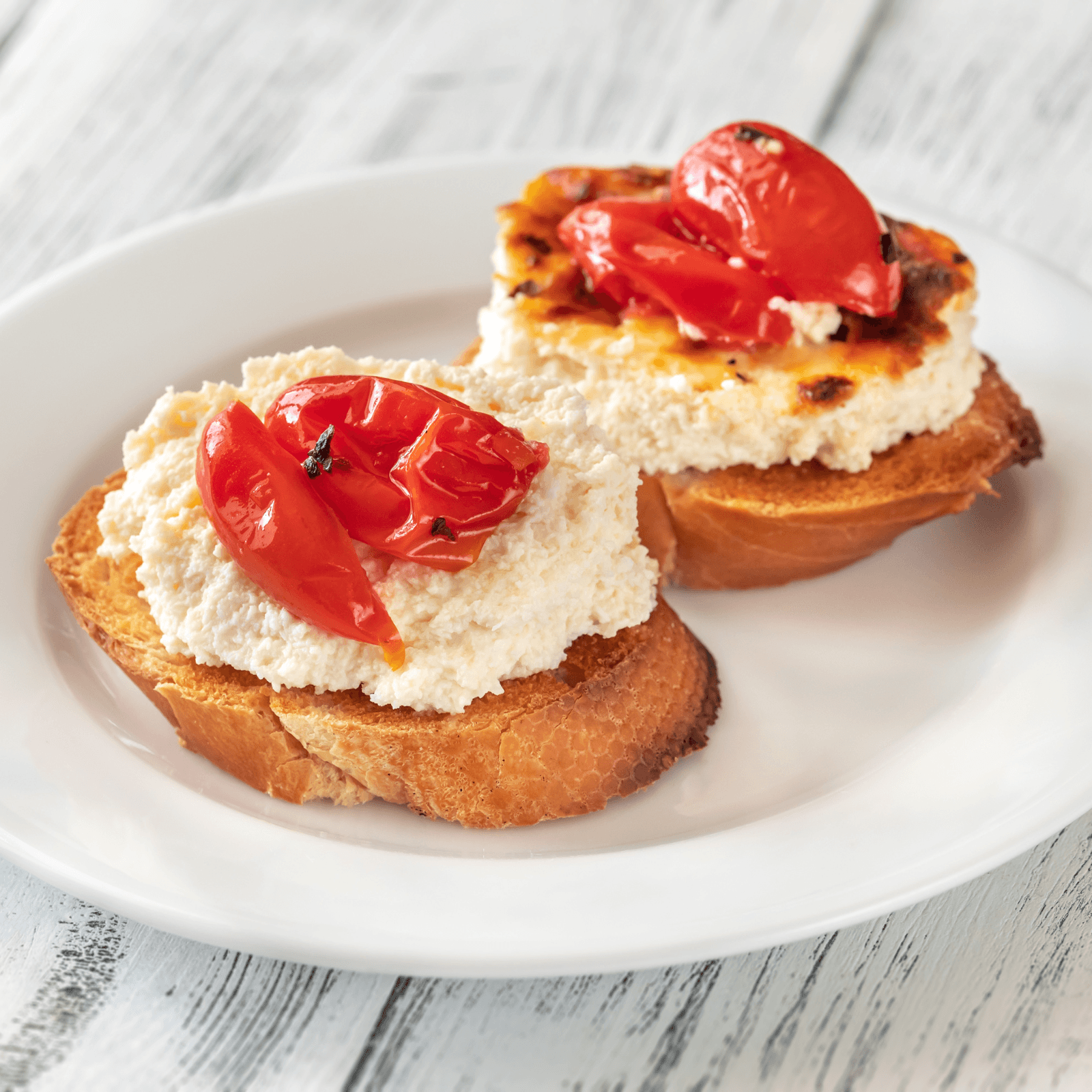 Image of two bruschetta appetizers with creamy ricotta cheese and roasted cherry tomatoes, served on a white plate with a light wooden background.
