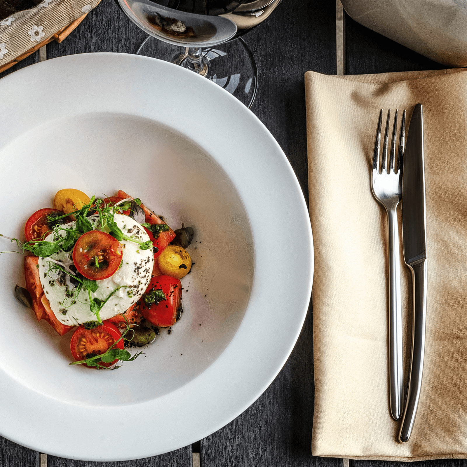 Overhead shot of a white plate containing a burrata salad with a creamy white burrata cheese ball, red and yellow cherry tomatoes, green herbs, and black pepper, positioned next to a beige napkin with a fork and knife, and the base of a wine glass, all on a dark wooden surface.