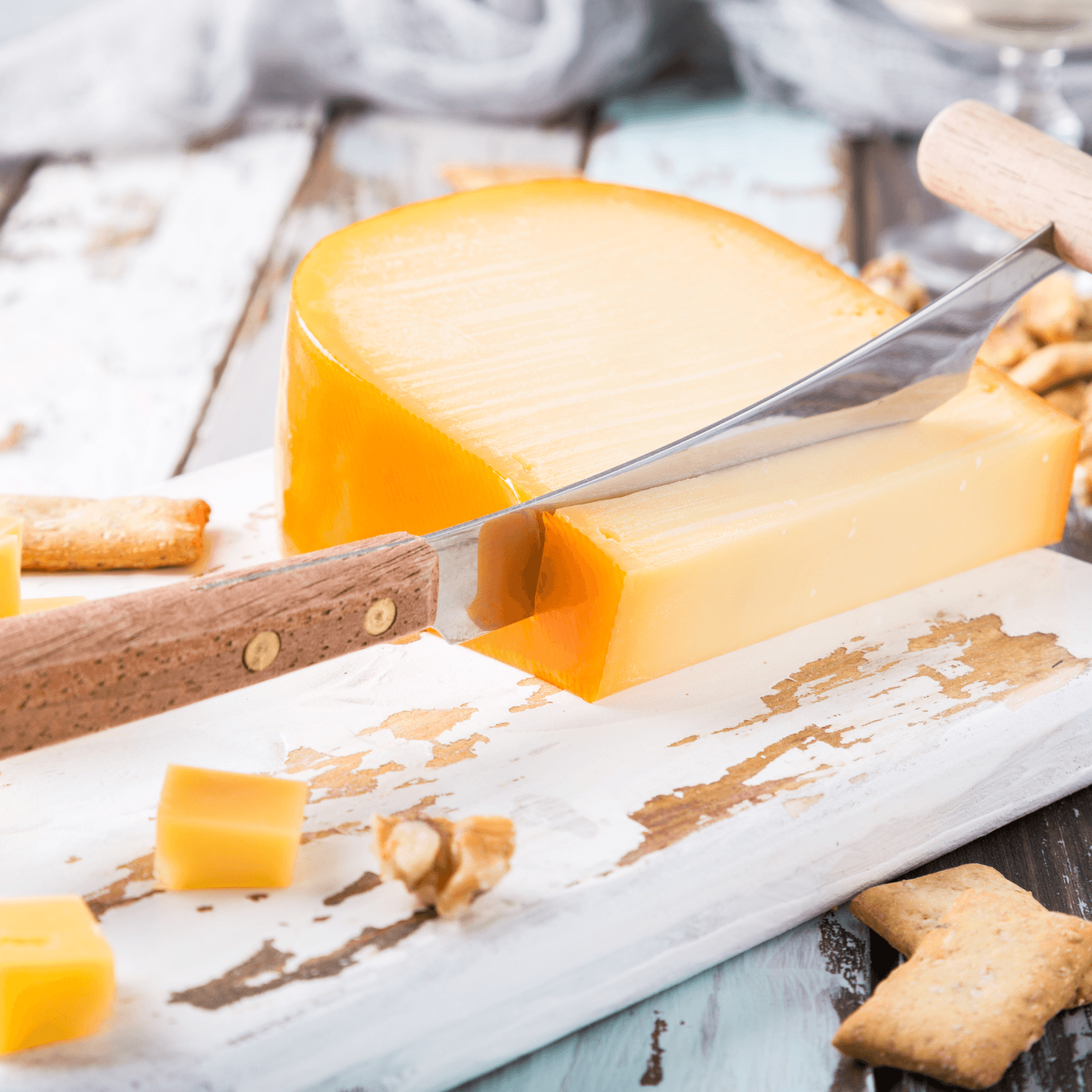 Close-up of Gouda cheese being cut with a knife on a white wooden serving board, showing its yellow color and texture, with crackers and walnuts as accompaniments.