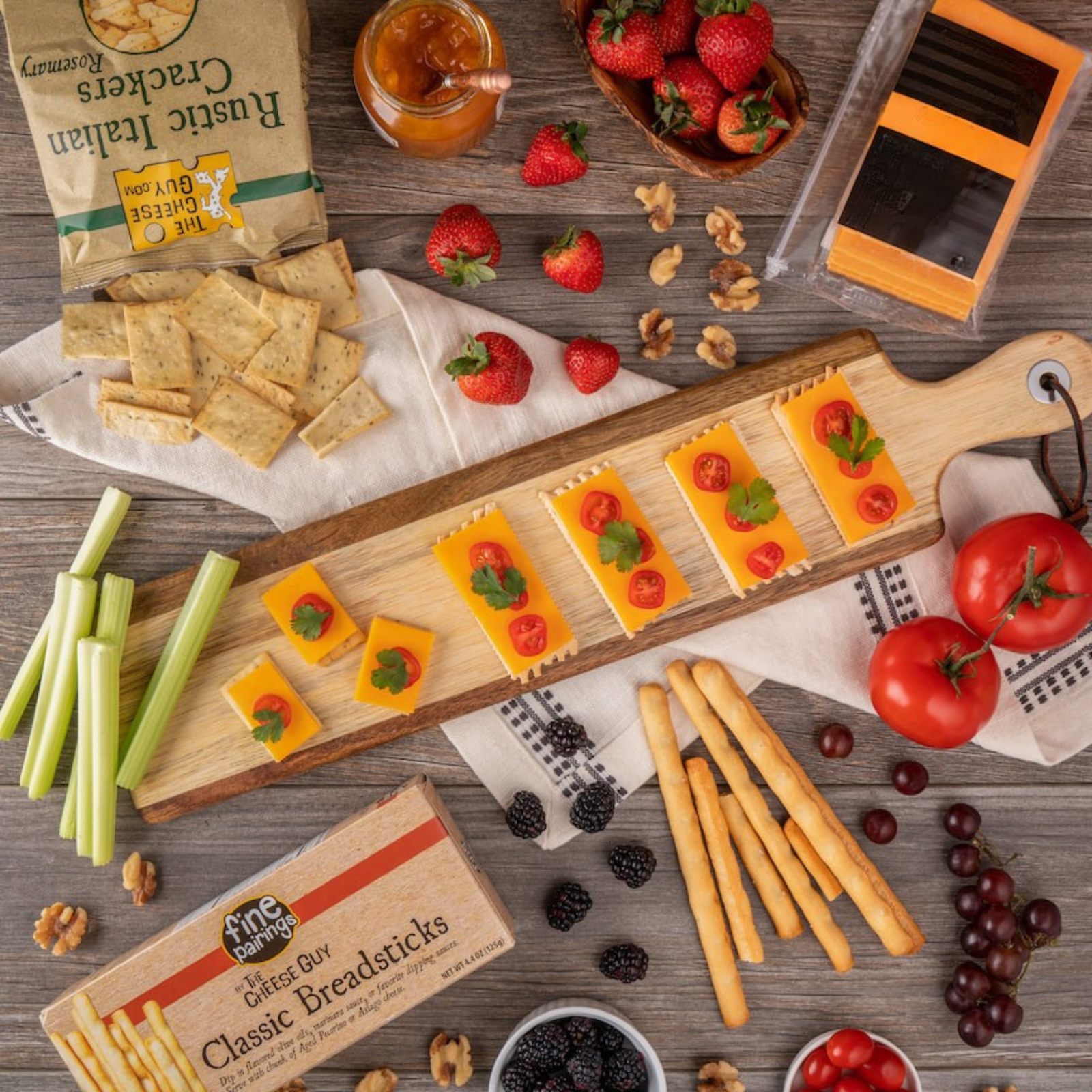 Artfully arranged overhead view of a cheese board with tomato and basil topped cheese slices, accompanied by a variety of complementary food items such as crackers, breadsticks, fresh berries (strawberries and blackberries), grapes, celery, nuts, and a jarred condiment.