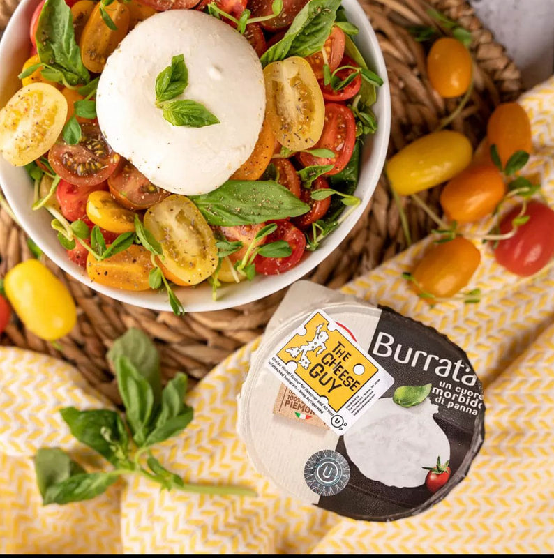 Overhead shot of a white bowl filled with a burrata salad featuring a creamy white burrata cheese ball topped with basil leaves, surrounded by colorful sliced cherry tomatoes and fresh basil, next to its packaging labeled "The Cheese Guy Burrata un cuore morbido di panna" on a yellow and white checkered surface with scattered cherry tomatoes.