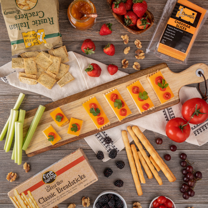Overhead view of a gourmet cheese board arrangement featuring The Cheese Guy Cheddar Slices topped with tomatoes and herbs on a wooden board, accompanied by The Cheese Guy Rustic Italian Rosemary Crackers, Fine Pairings Classic Breadsticks, celery sticks, fresh strawberries in a small basket, a jar of preserves with a spoon, whole tomatoes, blackberries, red grapes, and scattered walnuts, all on a rustic wooden surface with a white cloth napkin.