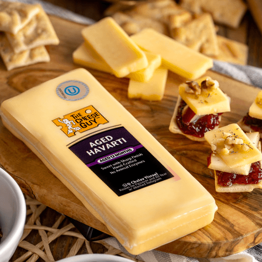 Overhead shot of a cheese board featuring a block of The Cheese Guy Aged Havarti with its purple label, several rectangular slices of Havarti, small crackers topped with cheese, a red spread, and nuts, along with scattered crackers, a white bowl filled with dark fruit (possibly prunes or figs), and a partially visible striped cloth napkin.