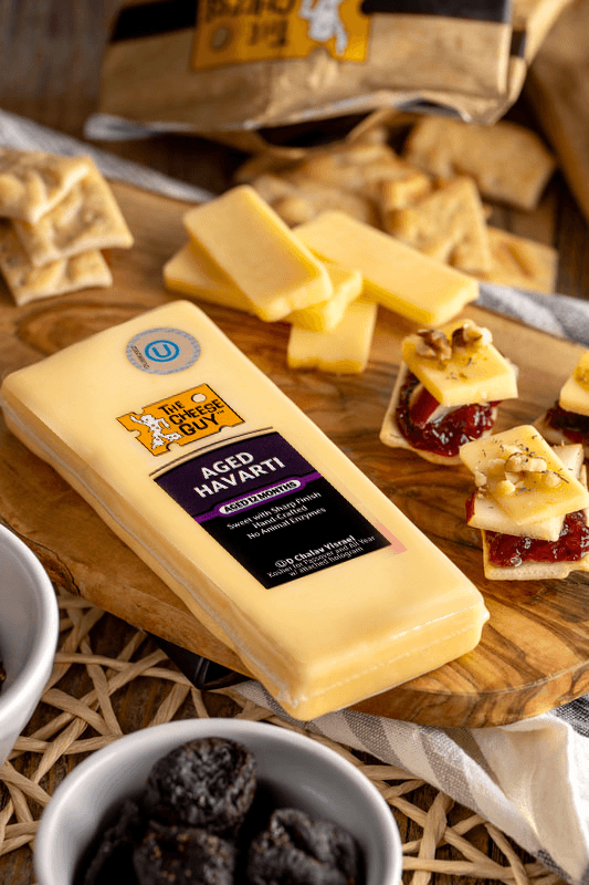 Overhead shot of a cheese board featuring a block of The Cheese Guy Aged Havarti with its purple label, several rectangular slices of Havarti, small crackers topped with cheese, a red spread, and nuts, along with scattered crackers, a white bowl filled with dark fruit (possibly prunes or figs), and a partially visible striped cloth napkin.