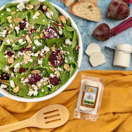Overhead shot of a spinach and beet salad in a white bowl featuring baby spinach, crumbled goat cheese, sliced roasted beets, and slivered almonds, accompanied by a package of The Cheese Guy Fresh & Creamy Goat Cheese with Honey, a wooden slotted spoon, roasted beets with their stems, slices of bread, and a log of goat cheese with two slices, all arranged on a light blue surface with a folded yellow cloth napkin.