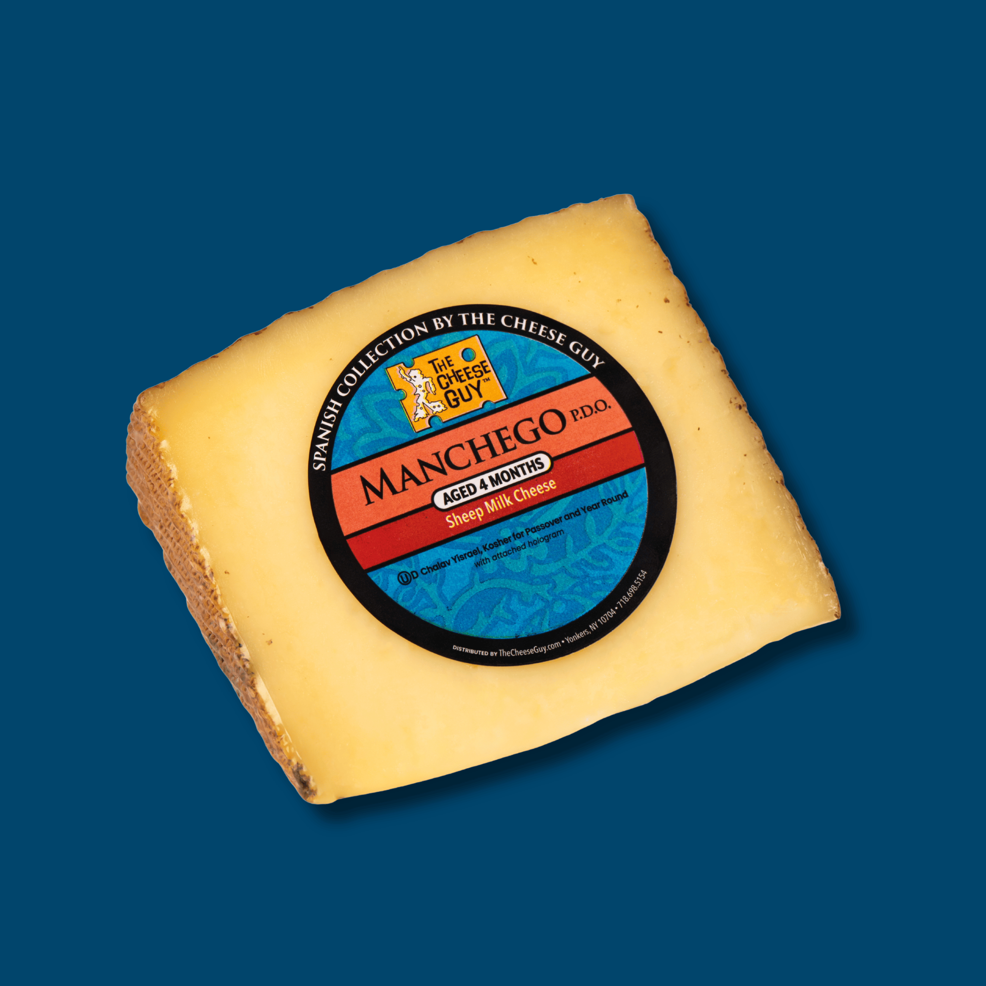 A wedge of The Cheese Guy Spanish Collection Manchego P.D.O. cheese, aged 4 months, made from sheep milk. The circular label states it's "Chalav Yisrael, Kosher for Passover and Year Round" and is prominently displayed on the rich yellow cheese on a blue background.