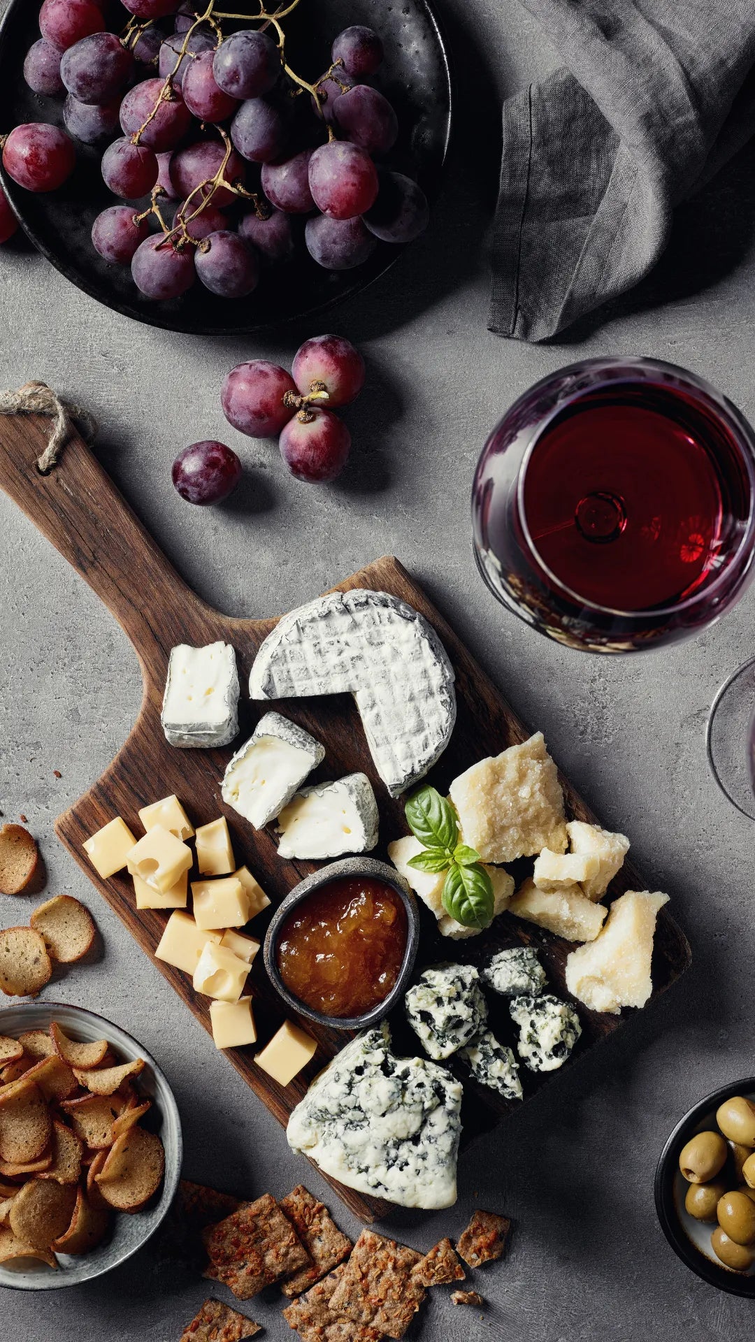 Top-down photo of a wooden serving board arranged with cheese (including white rind, yellow, and blue), grapes, crackers, jam, olives, nuts, and a glass of red wine on a textured gray background.