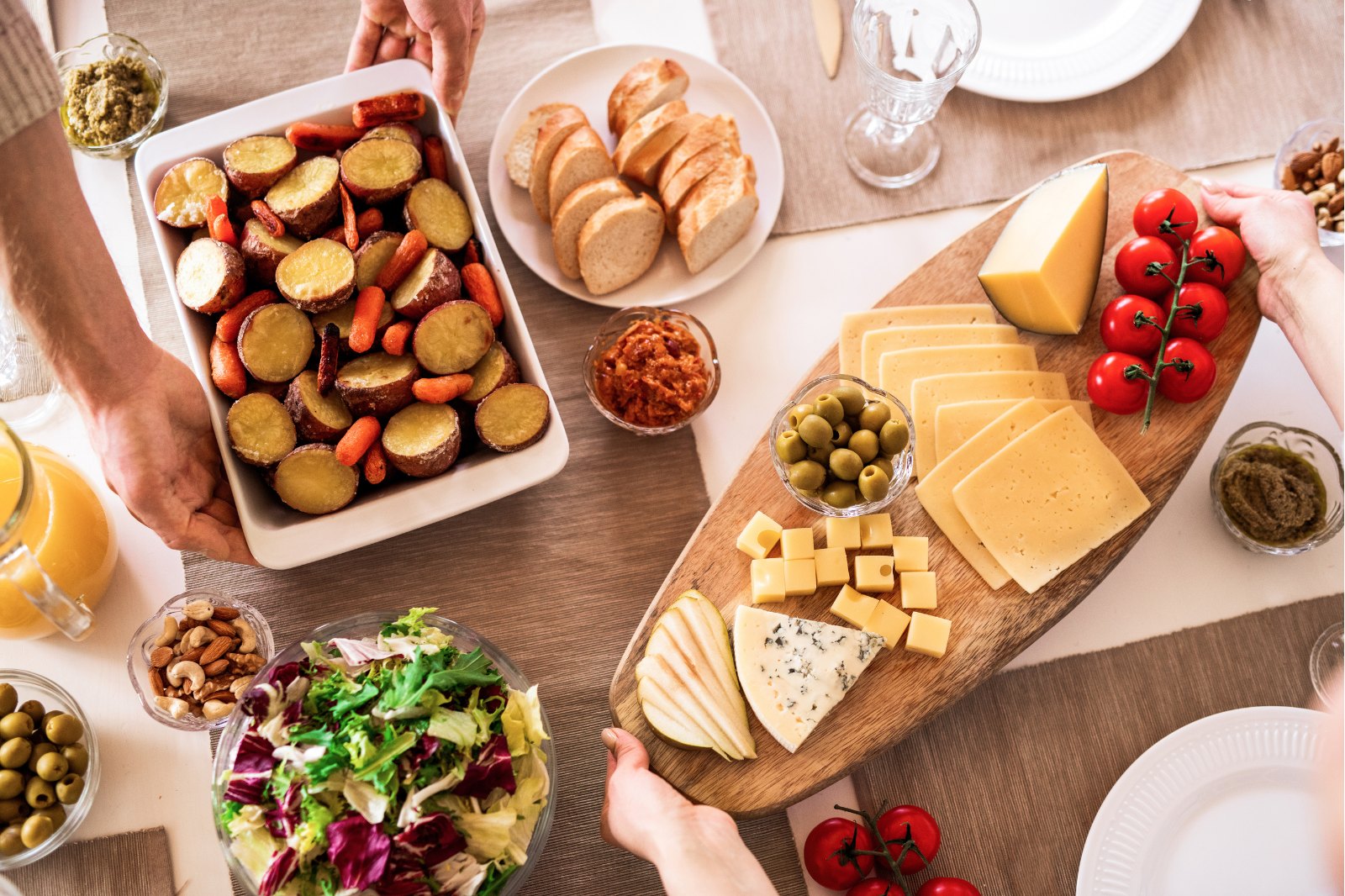 Top-down view of a table with hands reaching for various dishes. Roasted potatoes and carrots, sliced bread, cheese and tomato platter, salad, nuts, and dips are visible.