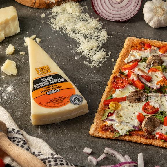 Overhead shot of ingredients for making pizza, featuring a wedge of The Cheese Guy Organic Pecorino Romano cheese with its label visible, a pile of shredded cheese, a partially visible block of cheese, sliced red onions, a whole white onion, and a rectangular flatbread pizza topped with sauce, cheese, mushrooms, red peppers, and red onions, all arranged on a dark grey surface with a wooden pizza peel handle and a white patterned cloth partially visible.