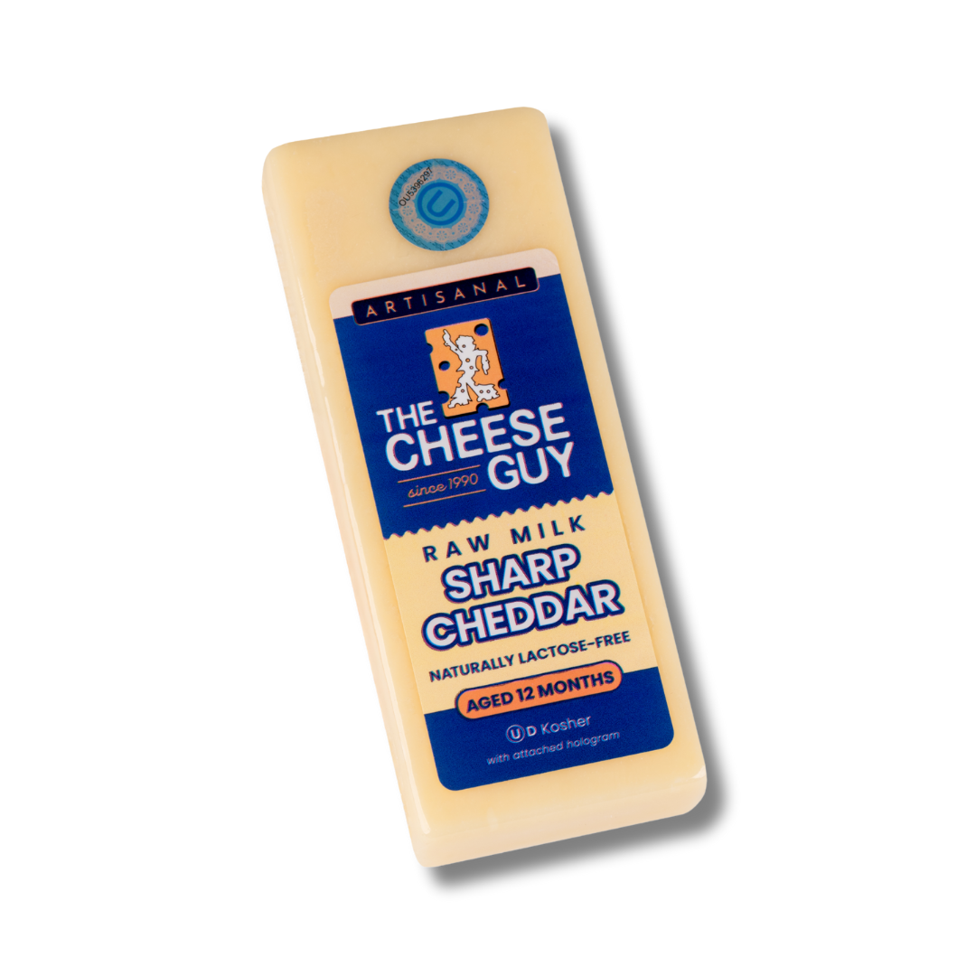 Image of The Cheese Guy Sharp Cheddar, a light-colored rectangular block of cheese, showing the brand logo, 'Sharp Cheddar' product name, and 'Aged 12 Months'.