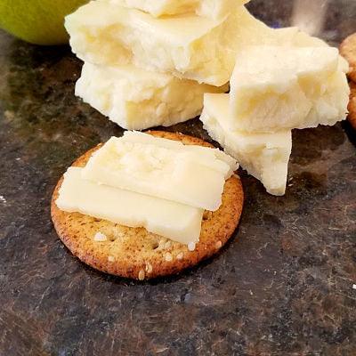 Close-up of a round cracker topped with slices of crumbly, pale white Wensleydale cheese, with a pile of similar cheese chunks in the background, all on a dark, speckled surface.