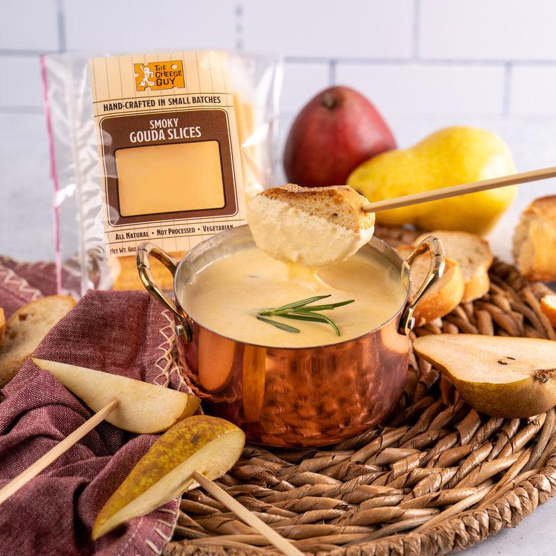 Image of cheese fondue featuring The Cheese Guy Smoky Gouda Slices, with bread dipped in the fondue, accompanied by pears, in a copper fondue pot on a woven tray.