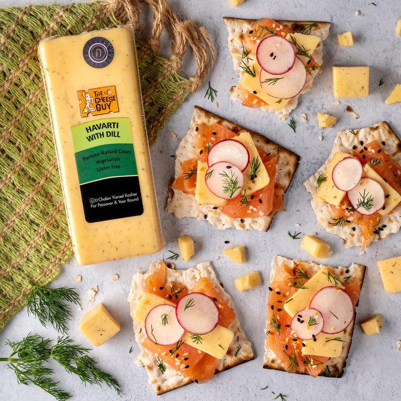 The Cheese Guy Havarti with Dill cheese, rectangular block, alongside crackers topped with salmon, cheese, and radish slices, with scattered cheese cubes and dill sprigs.