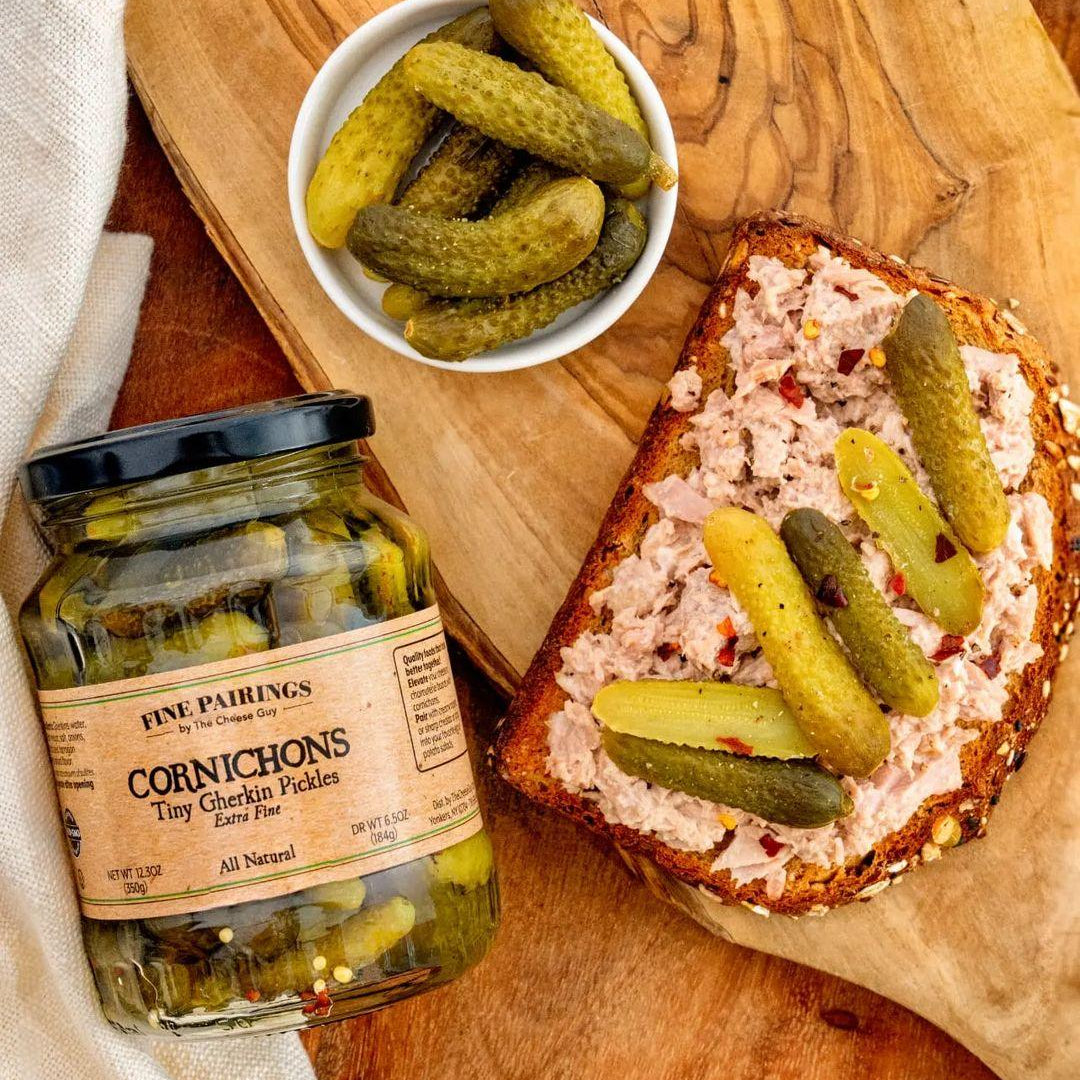 Image of a German Sausage Sandwich with liverwurst and Cornichons, served on a wooden board with a jar of Fine Pairings Cornichons and a small bowl of Cornichons, highlighting 'All Natural' on the label.
