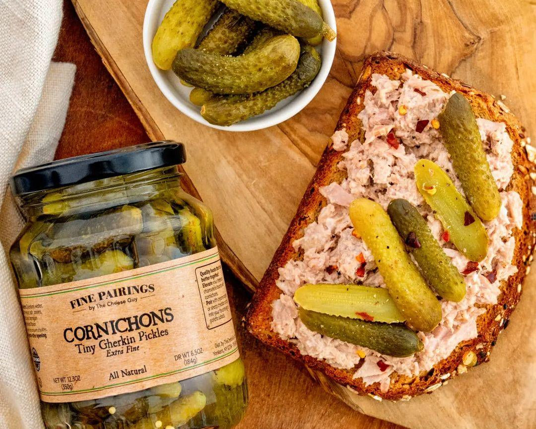 Image of a German Sausage Sandwich with liverwurst and Cornichons, served on a wooden board with a jar of Fine Pairings Cornichons and a small bowl of Cornichons, highlighting 'All Natural' on the label.